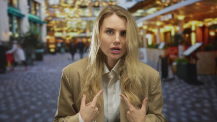 Fototapeta premium Woman gesturing on city street with relaxed expression, blonde hair, wearing beige jacket, surrounded by casual pedestrians and ambient, warm lighting in outdoor scene