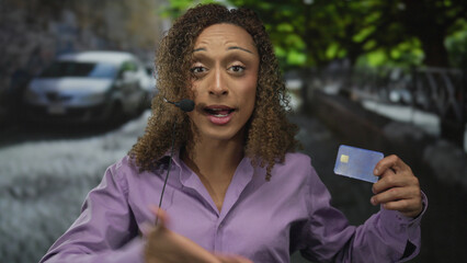 Woman holding blue creditcard and headset microphone on street pointing finger upward; confidence...