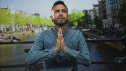 Young hispanic man with hands pressed together in praying gesture on a canal bridge street in amsterdam wearing a denim shirt and rings; hopeful prayer.
