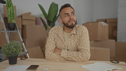Man with arms crossed amid cardboard moving boxes in a building interior of his new home, looking up with a pensive gaze; thoughtful planning.