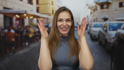 Fototapeta premium Woman frames face with hands while smiling on busy city street lined with parked cars and sidewalk cafes; playful joy.