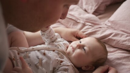 Adorable baby in onesie lying on bed, gazing at loving father leaning above her while bonding at home