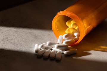 A dramatic, low-key photograph of capsules and pills spilling from an orange bottle onto a concrete surface, highlighted by hard light and deep shadow.