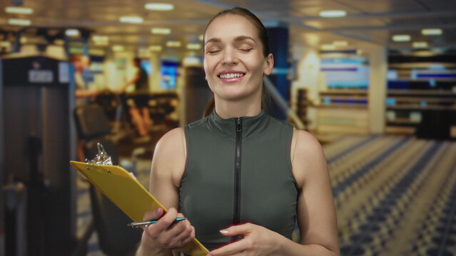 Woman with clipboard and pen gestures while discussing workout routine in a gym building; motivational support.