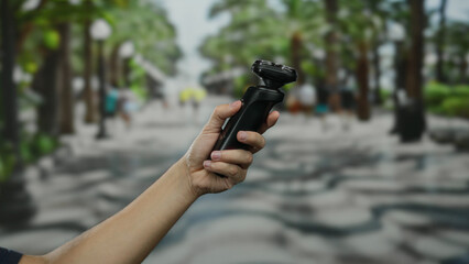 Man holding electric shaver in urban outdoor street setting with green trees and blurred background for modern lifestyle, grooming, and city life concept.