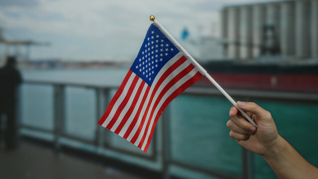 Man holding american flag at a seaside port, highlighting patriotism and maritime atmosphere, with a blurred boat backdrop creating a coastal setting.