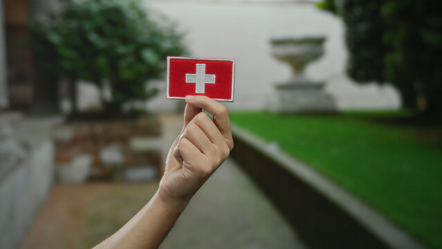 Man holding embroidered swiss flag patch with caucasian hand in a city outdoor setting against a blurred green background, symbolizing swiss culture and identity.