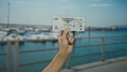 Hand holding united arab emirates dirham by the seaside with boats visible, highlighting the concept of finance in an outdoor maritime setting.