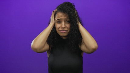 Woman with curly hair holding her head in frustration stands against a vivid purple background, expressing stress and concern in an isolated studio setting.
