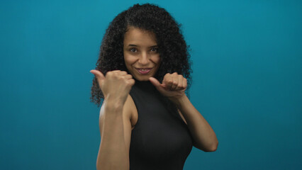 Woman with curly hair points her thumbs backwards with a playful smile in a blue studio; confidence...