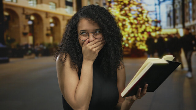 Young woman with glasses holding book on street with lights smiling with hand on mouth during evening
