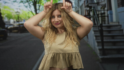 Woman blond young with bare arms points index fingers to her head on a street with stoop and lamppost, smiling at camera; playful mischief.