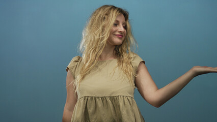 Fototapeta premium Woman raising arm with open palm and visible bare forearm, tousled blonde hair, relaxed pose in blue studio; playful warmth.