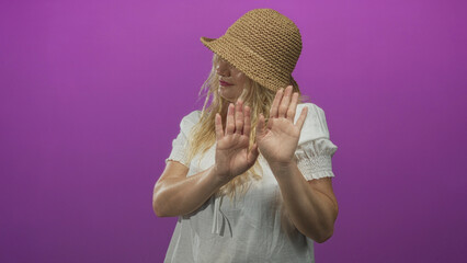 Woman blonde in a straw hat and white blouse holds hands up palms forward in purple studio with hat...