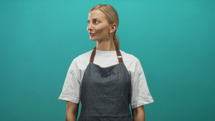 Woman in denim apron and white tshirt turns head to the side showing face and neck against teal studio backdrop; calm poise.