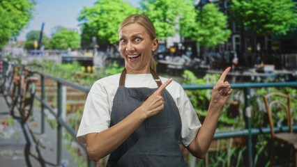 Fototapeta premium Blonde woman in apron points finger with both hands toward a canal railing on a busy street in daytime; playful surprise.