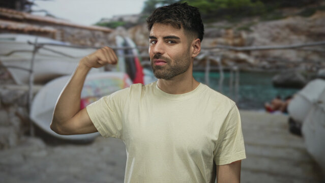 Man flexes bicep and stares at camera on a sandy beach near turquoise water under sunny sky; confidence. - Powered by Adobe