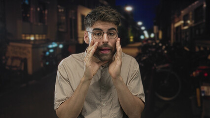 Young hispanic man with beard cups hands around mouth on dimly lit city street at night; urgent announcement.