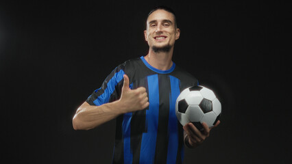 Man holding a soccer ball and points finger to ball in studio wearing blue striped jersey and smiling with thumbs up; confidence team spirit.