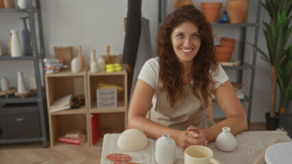 Woman artisan smiling with hands clasped over ceramic pots and clay on studio table wearing apron; craft focus serenity.