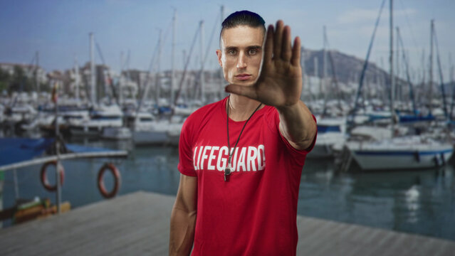 Man lifeguard in red shirt with whistle raising hand palm forward to stop on marina dock pier; vigilance duty. - Powered by Adobe
