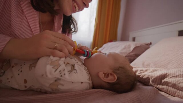 Young happy mother smiling as playing with baby lying on bed, grasping colorful rattle toy in sunlit room at home. Tilt-down shot