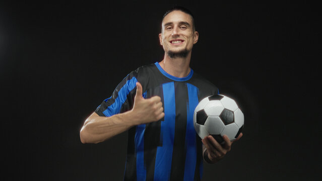 Man holding a soccer ball and points finger to ball in studio wearing blue striped jersey and smiling with thumbs up; confidence team spirit. - Powered by Adobe