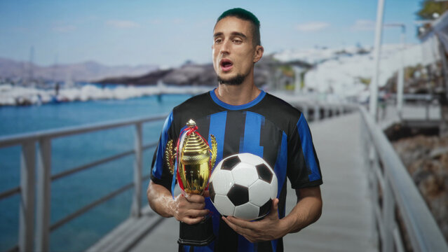 Man holding trophy and soccer ball, hands gripping both while eyes closed in blue black jersey on a street pier; victory pride.