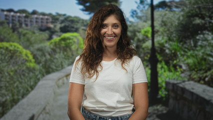 Woman smiling seated on a bench in forest setting wearing white t shirt and denim jeans, hands relaxed on lap; joy leisure nature.