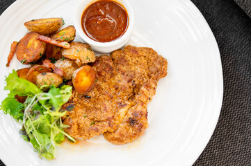 Crispy breaded pork cutlet served with roasted baby potatoes, a side salad, and dipping sauce on a white plate.