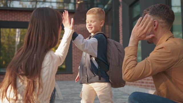 Caucasian boy running with backpack toward his happy parents near brick school building. Child smiling while rushing to greet them. Parents waiting with open arms ready to share warm moment.