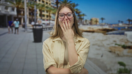 Fototapeta premium Young blonde woman wearing glasses laughs and covers mouth with hand on sunlit street by building; joy.