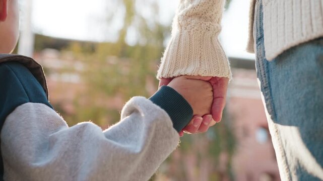 Close view child holding parent hand while standing together outside. Little boy gripping adult fingers with trust. Tender moment captured as they move along pathway before starting morning routine.