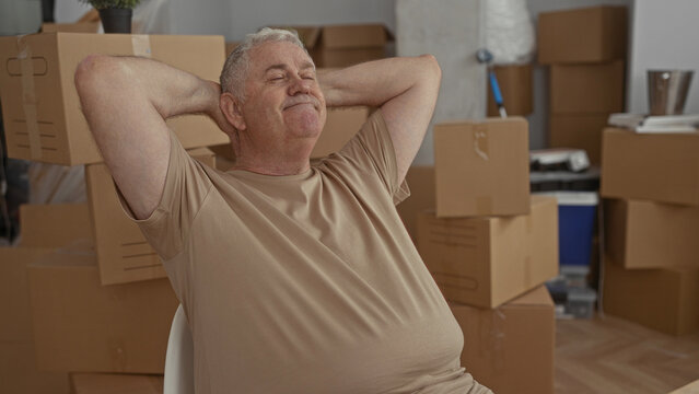 Man sitting on chair resting with hands behind head among stacked moving boxes in building interior, wearing beige tshirt and closed eyes; relief rest quiet.