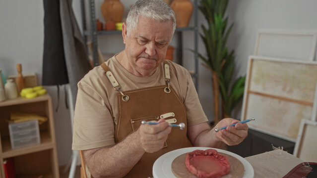Man in apron shaping clay on a turntable with modeling tools in studio, hands smoothing texture and forming a shallow bowl; craftsmanship patience.