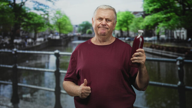 Man holding pomegranate juice bottle on a street bridge, examining label while smiling in maroon shirt; curiosity healthy choice.