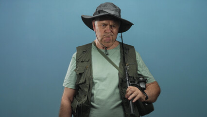 Man holds fishing rod and reel while wearing a boonie hat and fishing vest in studio; quiet...