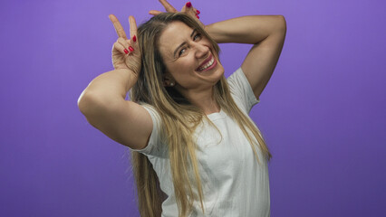 Middle aged blonde woman wearing white shirt smiling shows bunny ears gesture with hands in purple...