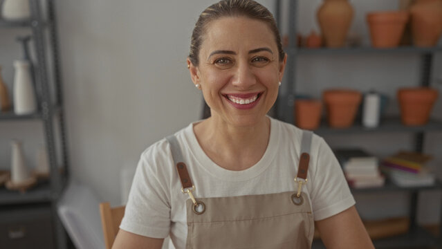 Artisan woman wearing apron smiles showing teeth beside ceramic pots and pottery shelves in studio workspace; joy.