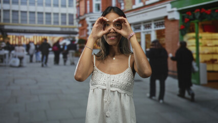Fototapeta premium Thai woman smiling with bare shoulders and hands forming binoculars on a busy city shopping street; playful joy.
