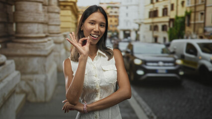 Fototapeta premium Thai woman shows ok hand gesture with fingers near face on a historic city street beside parked cars and stone building; playful travel.