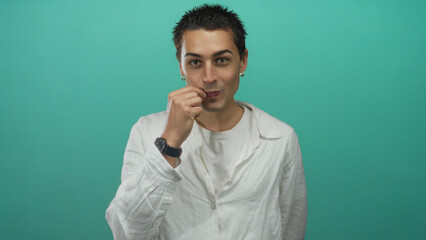 Fototapeta premium Young hispanic man making zip lips gesture in studio against green wall, wearing white long sleeve shirt and wristwatch; silence.