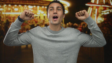 Young man enjoys lively outdoor night fair with blurred lights and crowd in background, capturing joyful atmosphere with confident expression and raised arms in casual sweater.