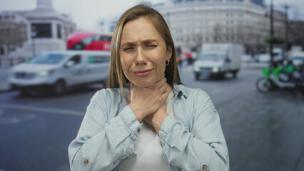Woman with sore throat standing on a busy urban street, expressing pain while traffic moves in the...