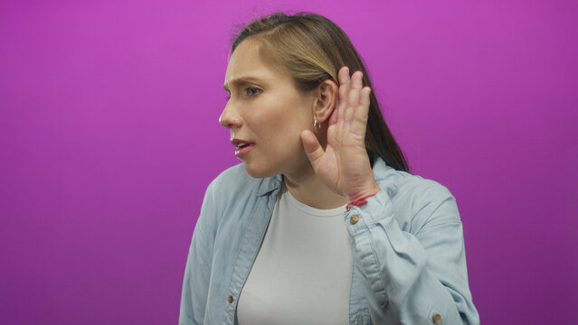 Woman listening intently with hand to ear against vibrant pink background in casual denim shirt suggesting attentive communication curiosity and engagement. - Powered by Adobe