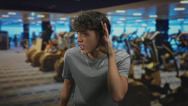 Teenage man cupping ear beside rows of treadmills, listening closely in a gym building; curiosity focus. - Powered by Adobe