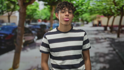 Teenage boy with glasses grinning and sticks out tongue, wearing a black and white striped tshirt while standing on a tree lined street with parked cars; playful.