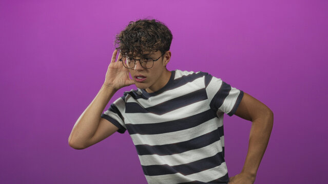 Teen boy with hand to ear listening gesture in studio purple backdrop, striped tshirt and glasses; curiosity confusion.