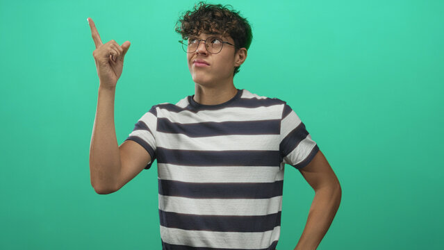 Teenage boy gestures thumbs down and points with fingers in a studio green screen set, wearing striped shirt and glasses; annoyance.