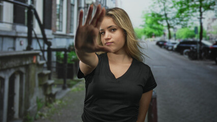 Fototapeta premium Woman young blonde palm forward stop gesture on narrow city street by brick building and parked cars; personal boundary defiance.